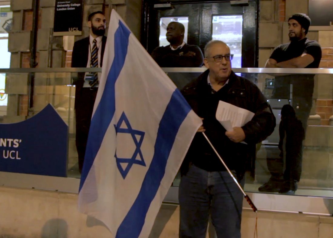 A man holding an Israeli flag stands outside of the Students' Union UCL building at 25 Gordon Street. Behind him are three security guards, looking on.