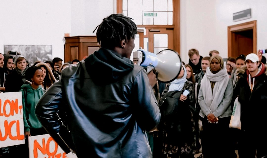 SU UCL BME Officer Ayo Olatunji, facing away from the camera, addresses a crowd of protesters using a megaphone at a protest against institutional racism.