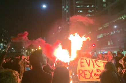 Student protestors carry a burning effigy of Rex Knight around UCL's Bloomsbury campus. There are red flares and a banner calling to evict Rex.